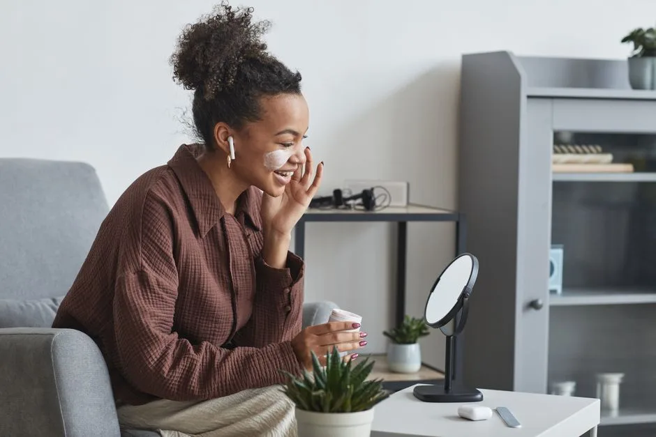 A woman applies facial cream while smiling indoors, capturing a moment of self-care using skincare products.
