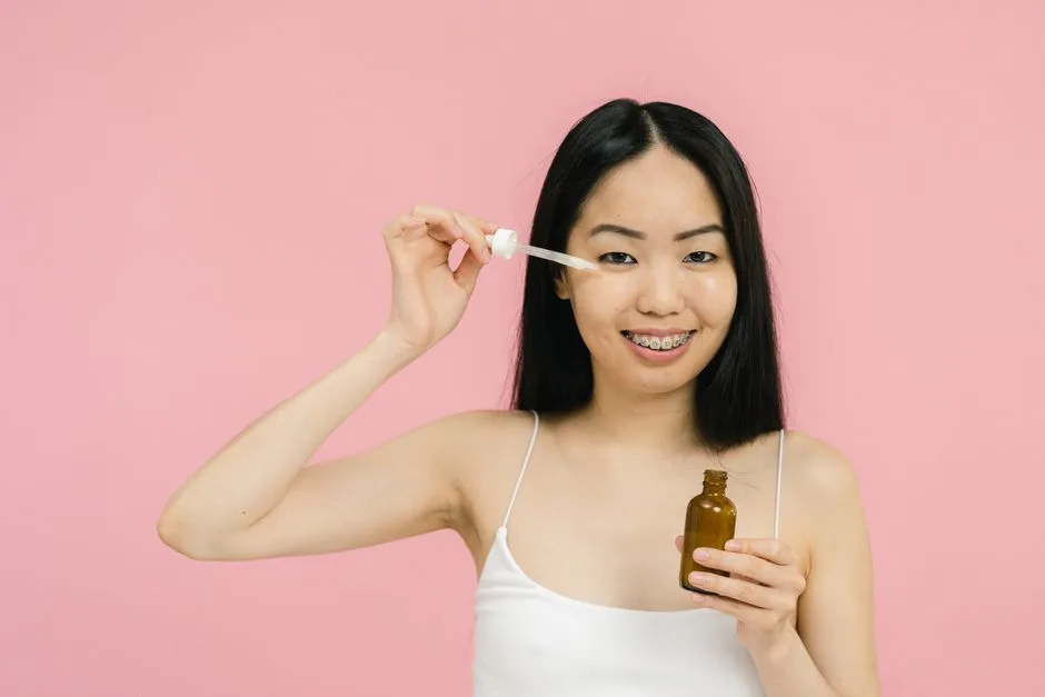Smiling woman with braces applying facial serum on a pink background, highlighting skincare routine.
